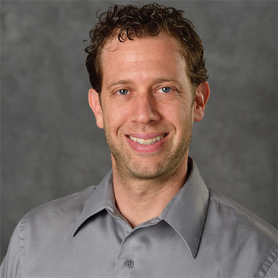 A headshot of Bryan Smith smiling wearing a gray button up