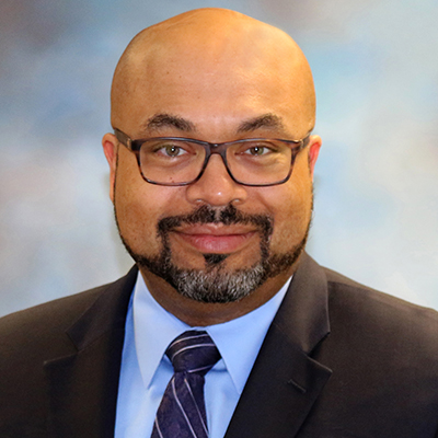 A headshot of Craig Cole smiling while wearing a black suit with a blue dress shirt and glasses