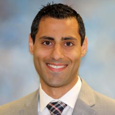 A headshot of Daniel Issac smiling wearing a tan blazer over a white dress shirt and plaid tie