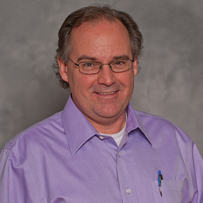 A headshot of Henry William smiling wearing glasses and a purple dress shirt