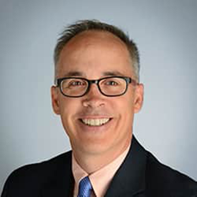 A headshot of Jeff Searl smiling wearing glasses and a black blazer over a pink dress shirt and blue tie