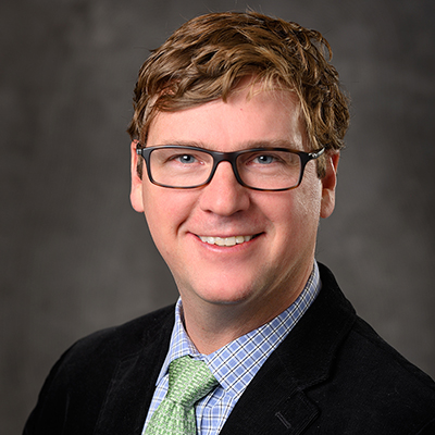A headshot of Jeffrey MacKeigan smiling wearing glasses, a black blazer over a blue dress shirt with a green tie