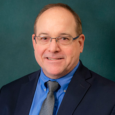 A headshot of Mark DeLano smiling while wearing a black suit and tie, blue dress shirt, and glasses