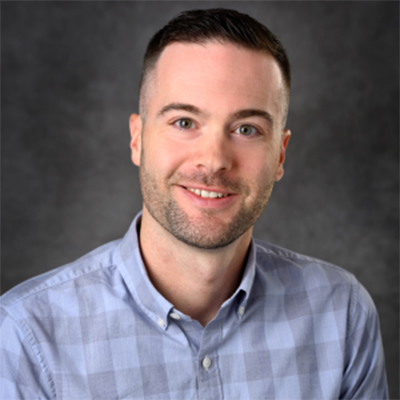 A headshot of Matthew Bernard smiling wearing a blue and gray plaid button up