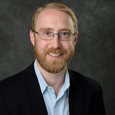 A headshot of Richard Lunt smiling wearing glasses with a black blazer over a blue dress shirt