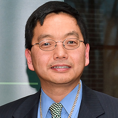 A headshot of Zuefei Huang smiling wearing glasses with a black blazer of a blue dress shirt and green tie