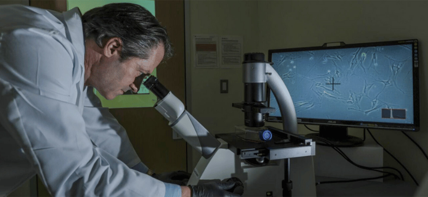 A photo of André Bachmann wearing a lab coat and looking into a microscope in a lab
