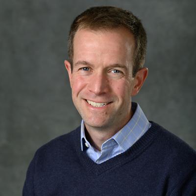 A headshot of Adam Alessio smiling with a blue sweater over a polo