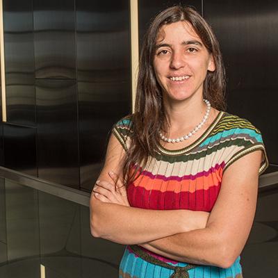A headshot of Ana Vazquez crossing arms smiling wearing a colorful top and pearl necklace