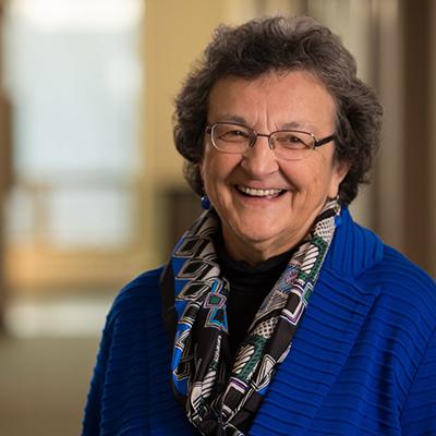 A headshot of Barbara Given with glasses smiling while wearing a blue sweater and multi-colored scarf