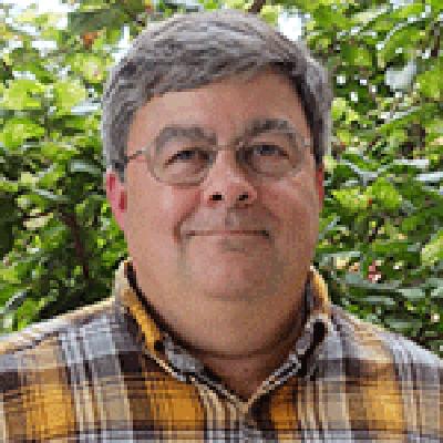 A headshot of Brad Upham wearing glasses smiling in a yellow plaid button up