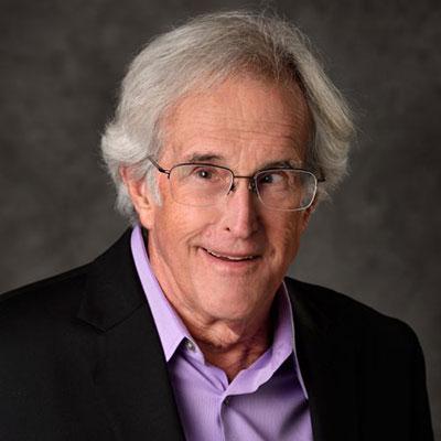 A headshot of Charles Given wearing glasses smiling while wearing a black blazer and purple dress shirt