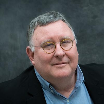A headshot of Edmund Ellsworth smiling in a black blazer over a blue dress shirt and wearing glasses