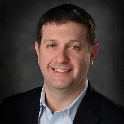 A headshot of Erik Shapiro smiling wearing a black blazer over a blue dress shirt