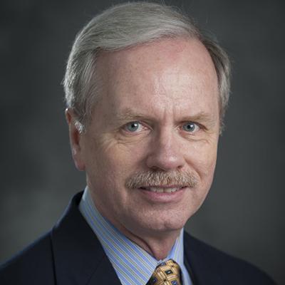 A headshot of Gale Strasburg smiling wearing a black blazer over a blue and gray striped button up with a yellow tie