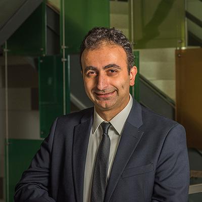 A headshot of George Mias smiling in a black blazer and tie over a white dress shirt