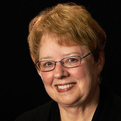 A headshot of Gwen Wyatt smiling wearing glasses and a black blazer