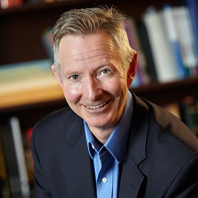 A headshot of James Dearing smiling while wearing a black suit over a blue button up in front of a bookshelf