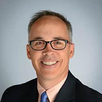 A headshot of Jeff Searl smiling wearing glasses and a black blazer over a pink dress shirt and blue tie