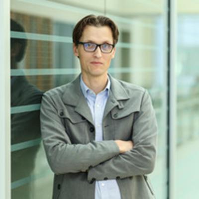 A headshot of Jeffrey Klomp wearing glasses crossing his arms wearing a gray blazer with a blue dress shirt underneath