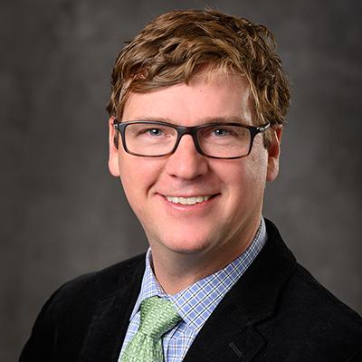 A headshot of Jeffrey MacKeigan smiling wearing glasses, a black blazer over a blue dress shirt with a green tie