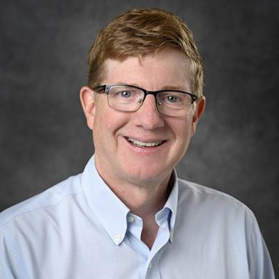 A headshot of Kurt Zinn smiling wearing glasses and a blue dress shirt
