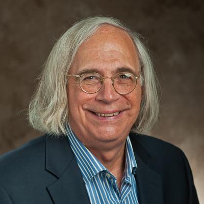 A headshot of Leonard Fleck smiling with glasses while wearing a black blazer with a blue and white striped dress shirt underneath. 