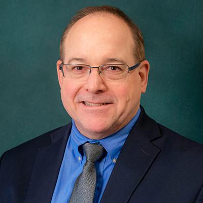 A headshot of Mark DeLano smiling while wearing a black suit and tie, blue dress shirt, and glasses