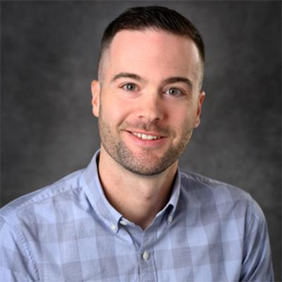 A headshot of Matthew Bernard smiling wearing a blue and gray plaid button up