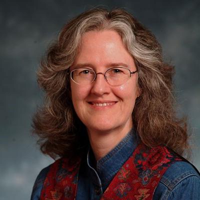 A headshot of Nancy Turner wearing glasses smiling while wearing a jean top and red scarf