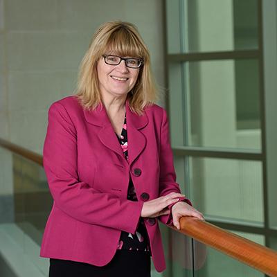 A headshot of Rebecca Lehto smiling with glasses wearing a pink blazer and posing near a railing