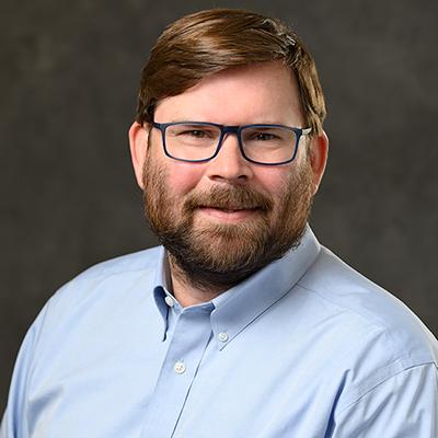 A headshot of Ron Chandler smiling while wearing a blue dress shirt and glasses