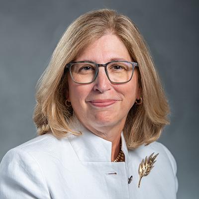 A headshot of Teresa Woodruff smiling wearing glasses and a white top with a gold badge, necklace, and earrings