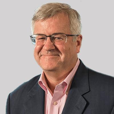 A headshot of Thomas O'Halloran smiling wearing glasses with a black blazer and pink dress shirt