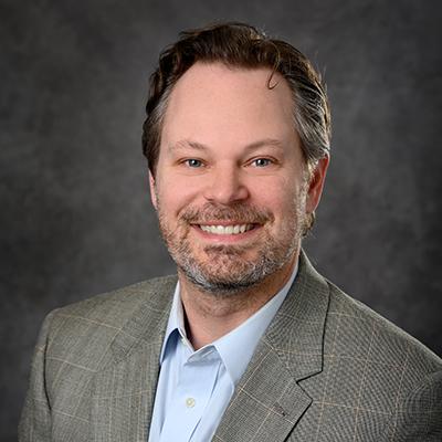 A headshot of Todd Lucas smiling wearing a gray blazer over a blue dress shirt