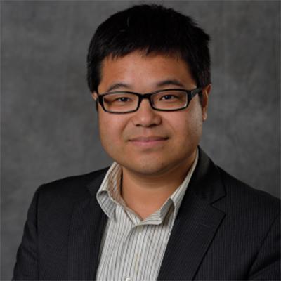 A headshot of Zhen Qiu smiling wearing glasses and a black blazer over a striped dress shirt